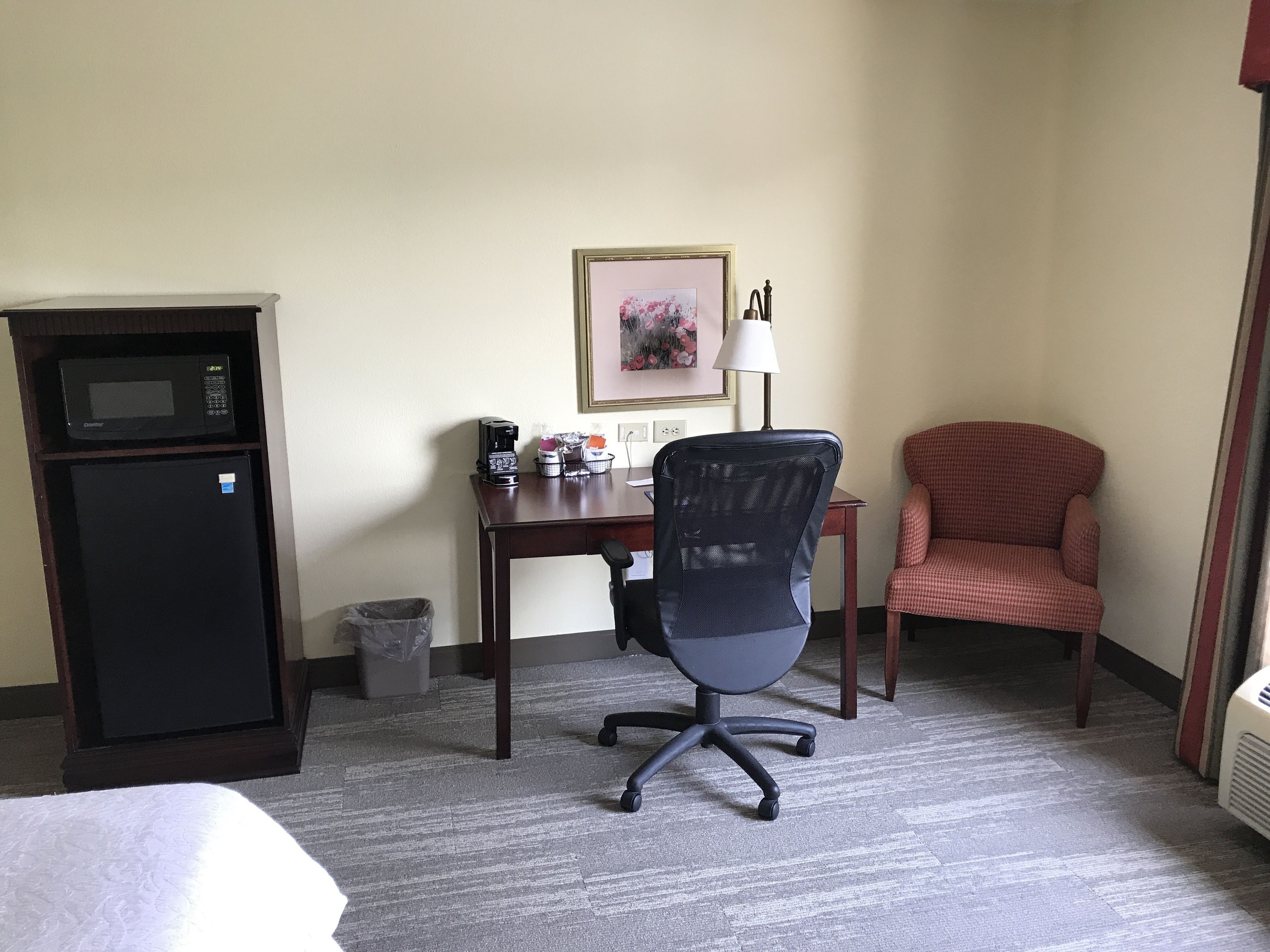 A hotel room with a bed, a desk with a computer, a chair, a microwave, a trash can, and a picture on the wall.
