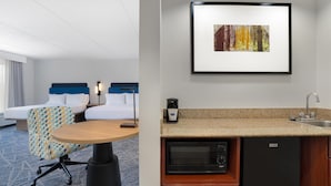 Habitación de hotel con cama, escritorio, silla, microondas, cafetera y un cuadro en la pared.