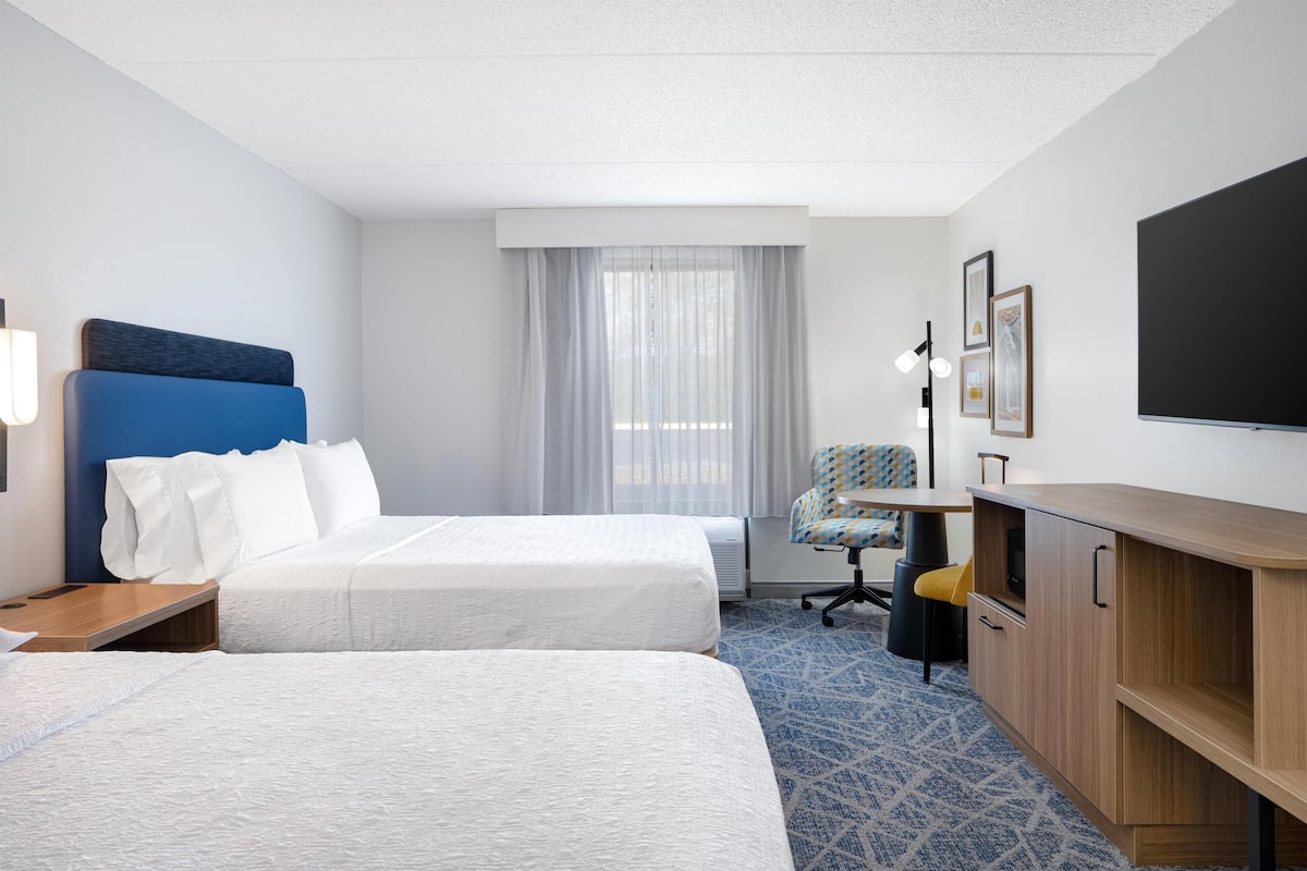 A hotel room with two beds, a desk, a chair, a TV, and a large window with curtains.