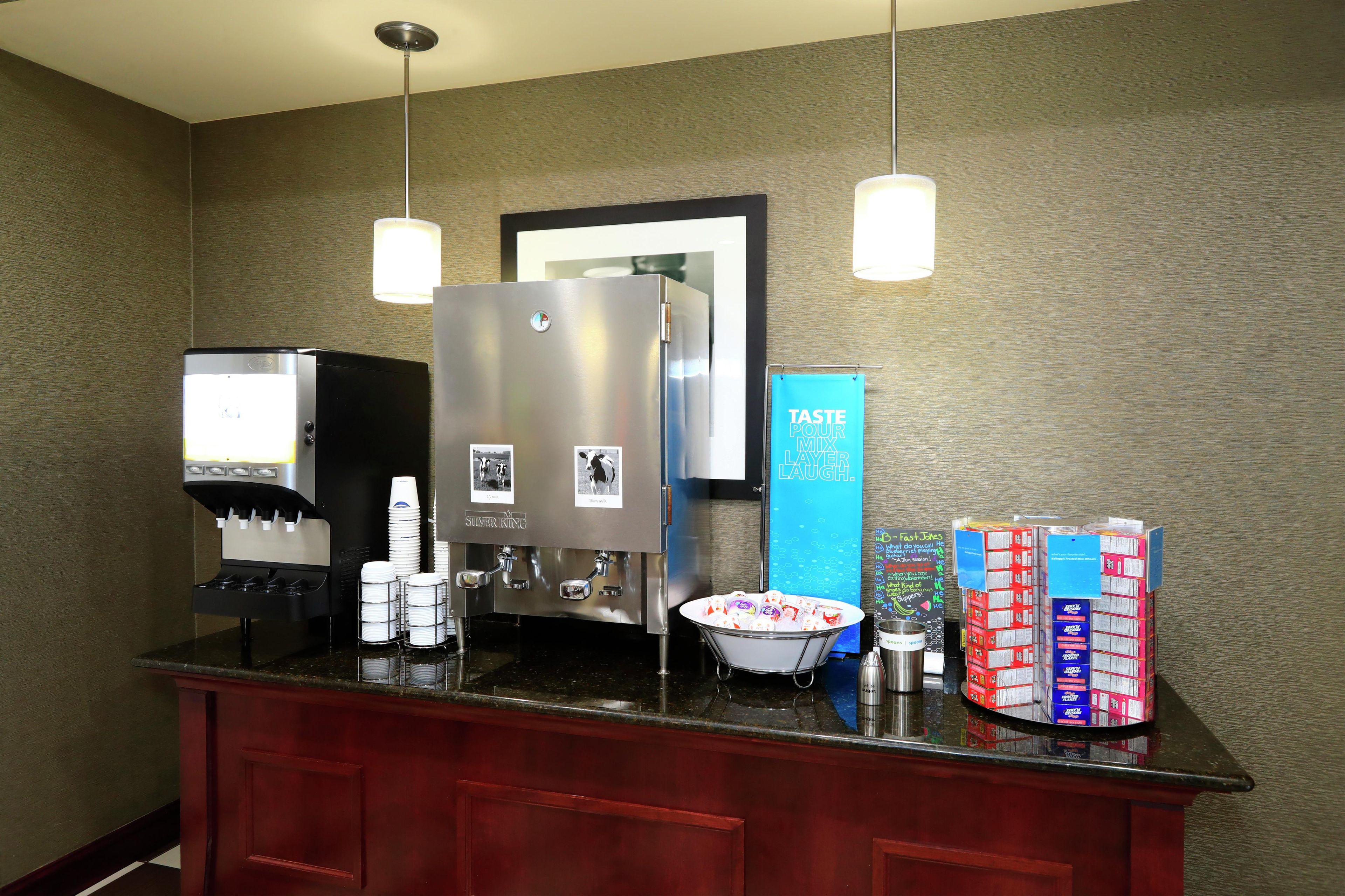 Zona de recepción de un hotel con una máquina de café, un refrigerador, un cuenco de bocadillos y un cartel que dice 'TASTE POUR MIX LAYER LAUGH'.