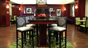 A hotel dining area with a central table surrounded by chairs, a television above the table, and framed pictures on the walls. - Hampton Inn & Suites Lamar (Mill Hall)