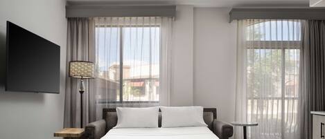 A modern hotel room with a large bed, a flat-screen TV, a small table, and a balcony with curtains.
