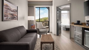 A modern hotel room with a grey sofa, a wooden coffee table, a grey armchair, a dining table with chairs, a kitchen area, and a balcony with a view of trees and buildings.