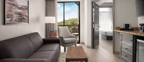 A modern hotel room with a grey sofa, a wooden coffee table, a grey armchair, a dining table with chairs, a kitchen area, and a balcony with a view of trees and buildings.