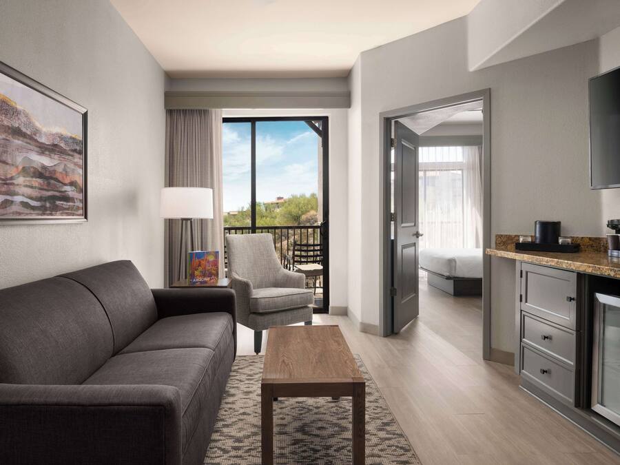 Une chambre d’hôtel moderne avec un canapé gris, une table basse en bois, un fauteuil gris, une table à manger avec des chaises, un coin cuisine et un balcon offrant une vue sur les arbres et les bâtiments.
