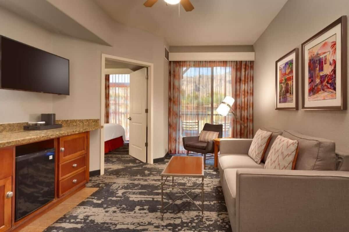 Une chambre d’hôtel équipée d’un téléviseur à écran plat, d’une kitchenette, d’un canapé, d’une table basse et d’une grande fenêtre avec des rideaux.
