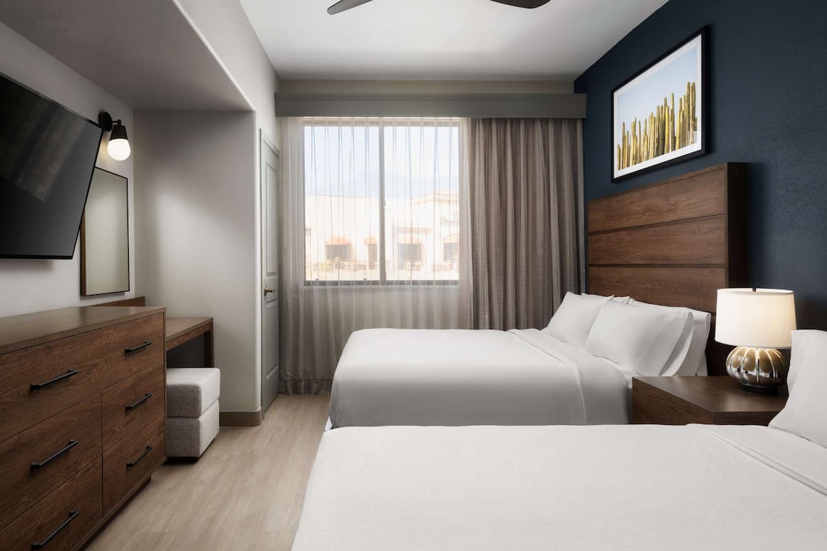 Une chambre d’hôtel avec un lit, une table de chevet, une lampe, une commode, un miroir et une fenêtre avec des rideaux.