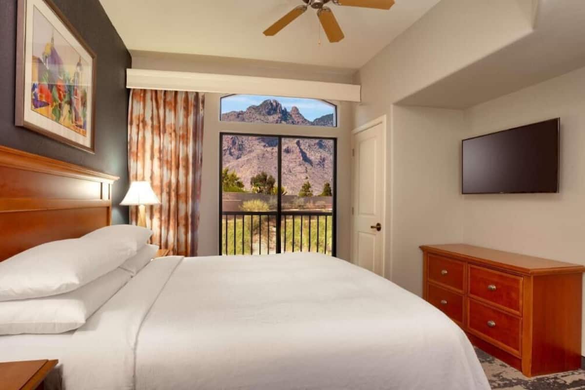 Une chambre d’hôtel comprenant un lit, une télévision, une commode en bois, un ventilateur de plafond, une fenêtre donnant sur la montagne et un tableau encadré au mur.