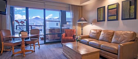 A modern hotel room with a wooden coffee table, leather sofa, and a dining area with chairs and a table. Large windows offer a view of mountains and a cityscape.