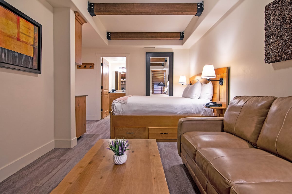 A bedroom with a bed, a wooden nightstand with a lamp, a brown leather sofa, a wooden coffee table with a plant, and a framed picture on the wall.