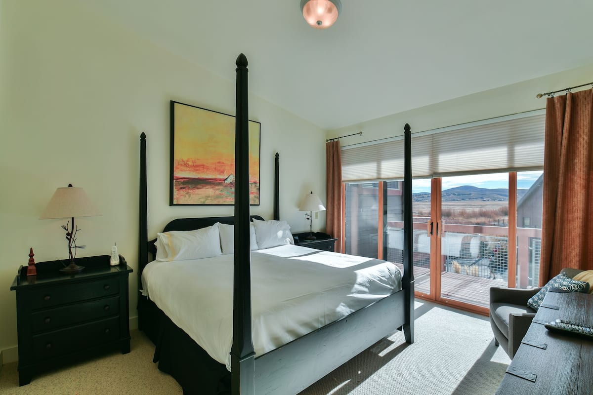 A four-poster bed with a city view, a nightstand with a lamp, and a window with blinds.