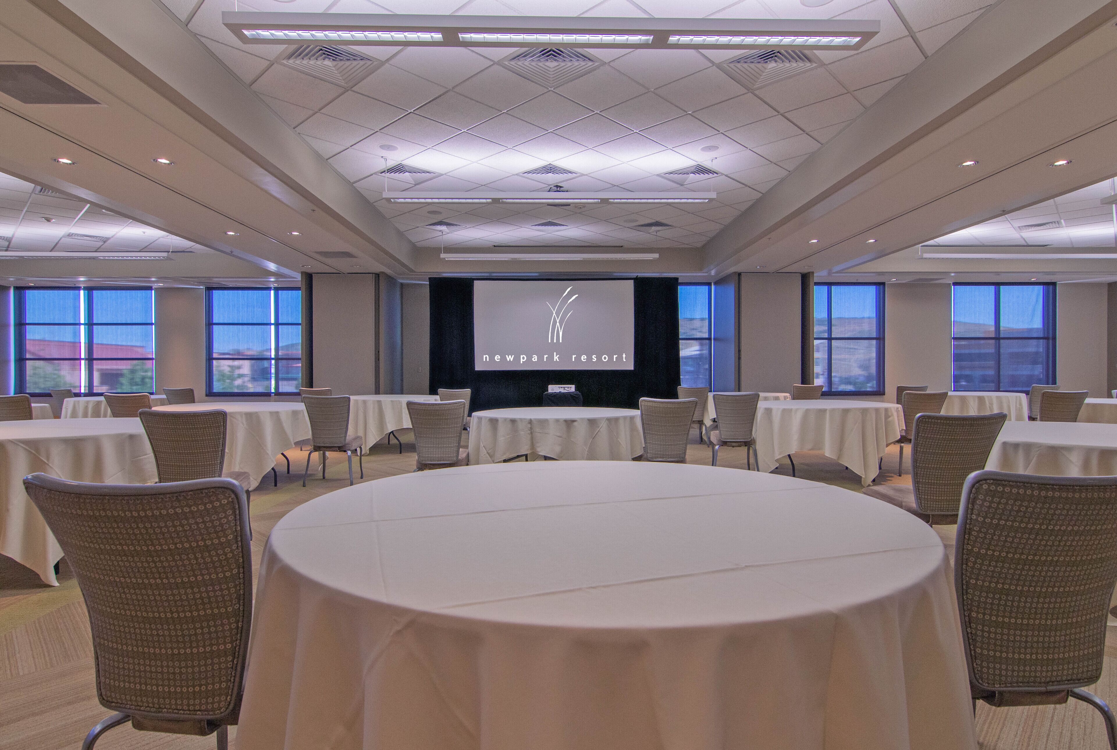 A conference room with round tables, chairs, a large screen, and a view of the outdoors.