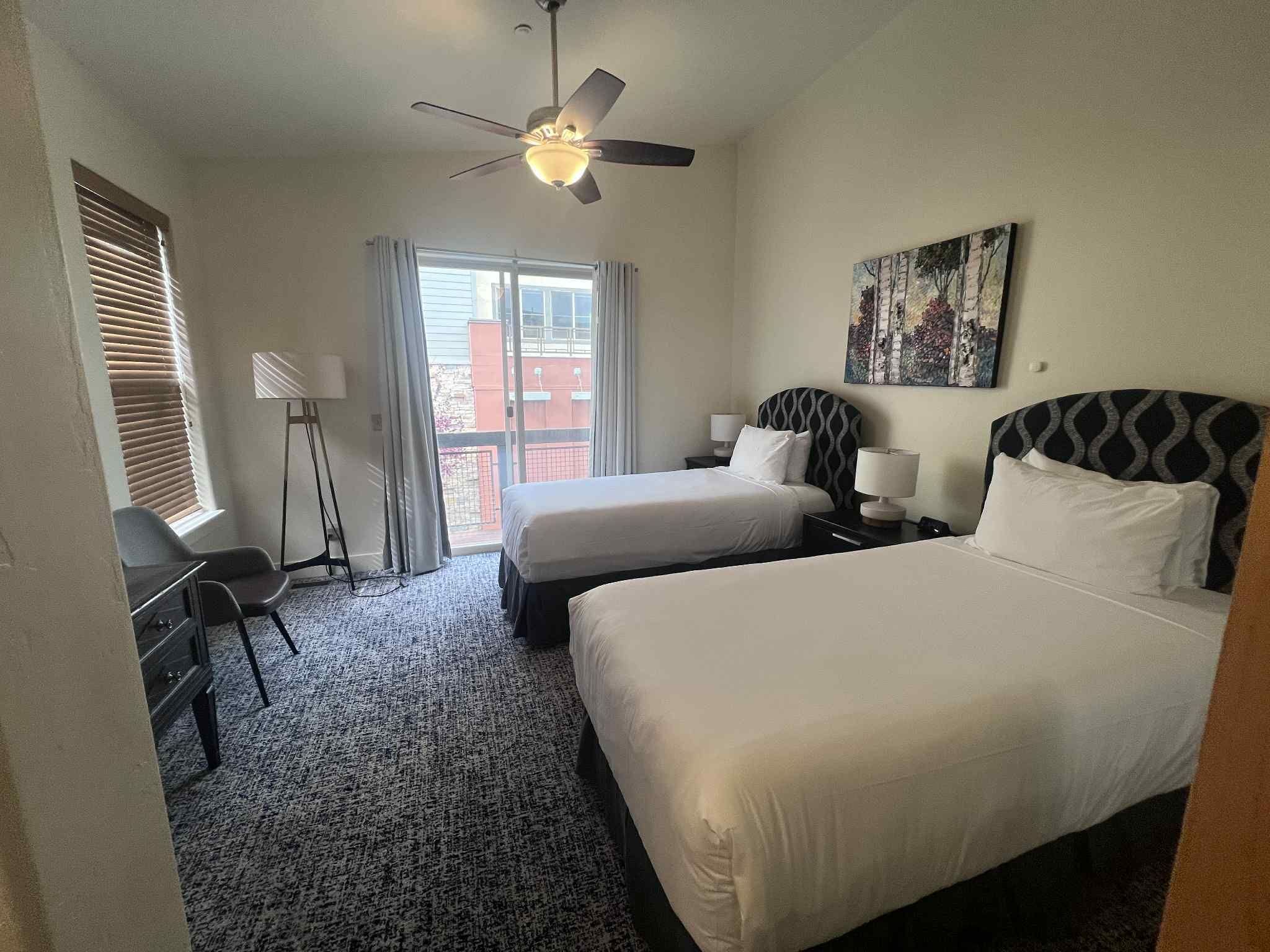 Une chambre d’hôtel avec deux lits, un bureau, une chaise, un ventilateur de plafond et une fenêtre avec des stores.