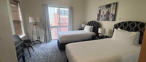 A hotel room with two beds, a desk, a chair, a ceiling fan, and a window with blinds.