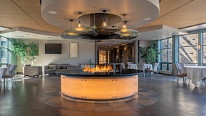 A modern lounge area with a central fire feature, seating arrangements, and large windows providing natural light. - Newpark Resort (Park City)