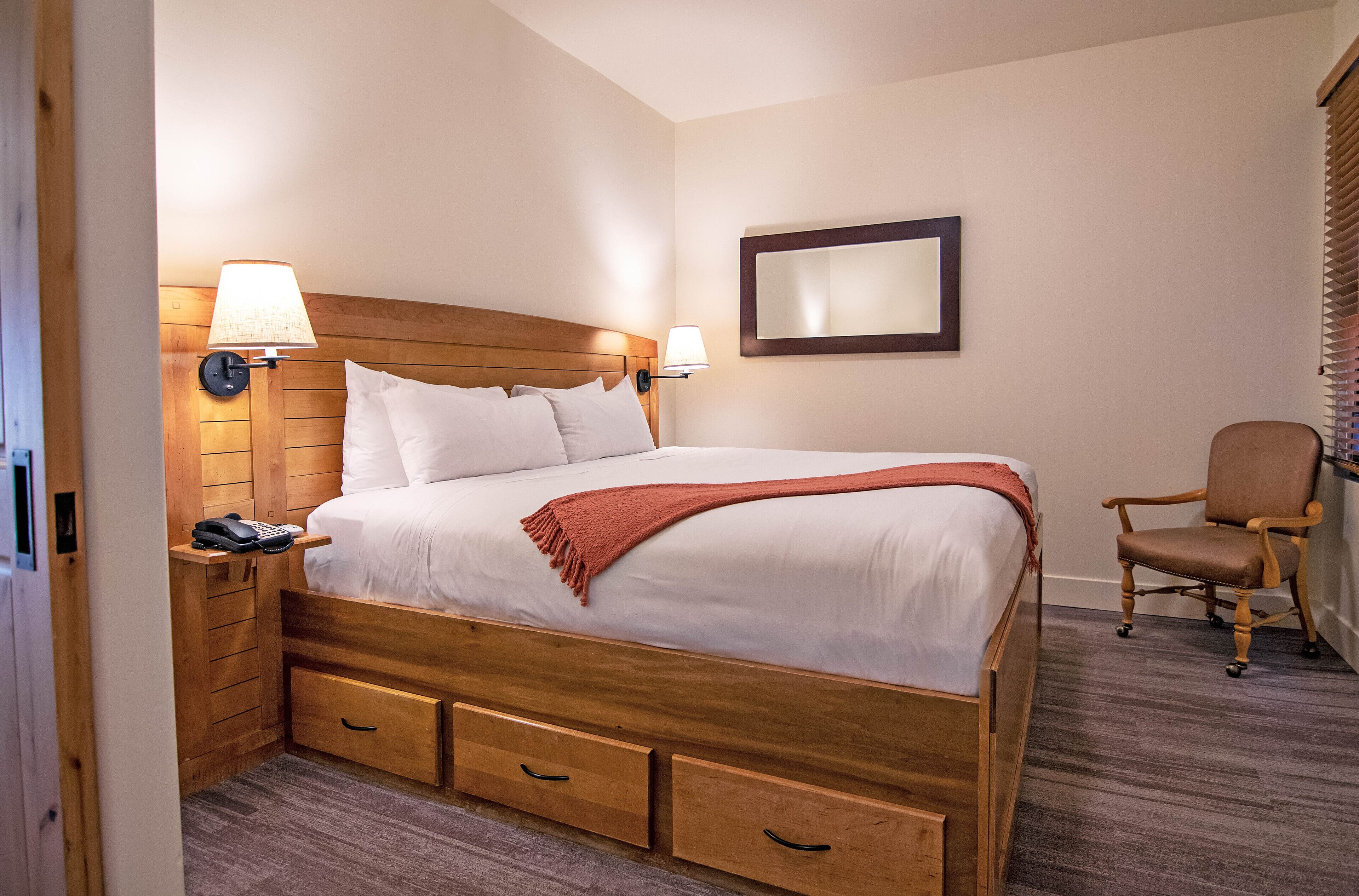 A hotel room with a wooden bed, bedside lamps, a desk with a phone, a chair, and a mirror on the wall.