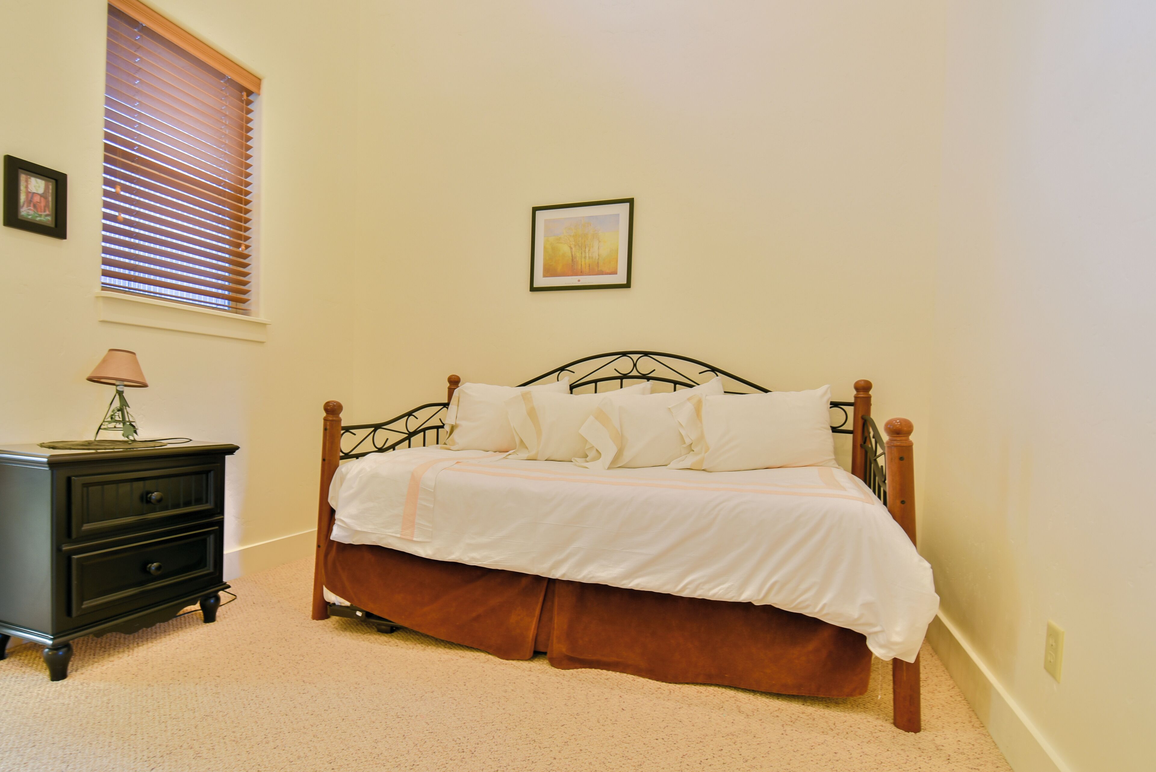 A bedroom with a bed, a nightstand, a lamp, a window with blinds, and a framed picture on the wall.