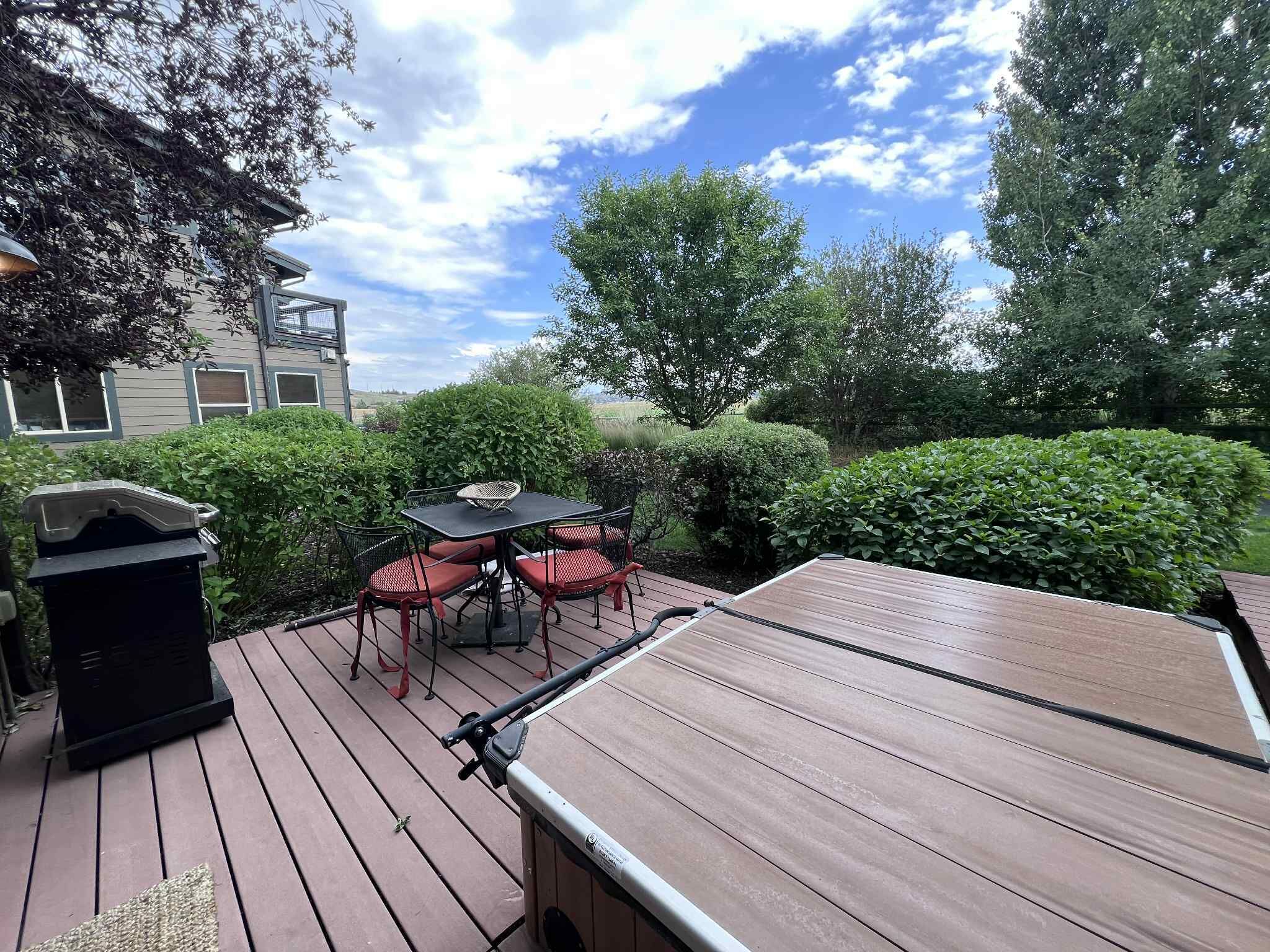 Una terraza de madera con una mesa y sillas, rodeada de follaje y un edificio al fondo.