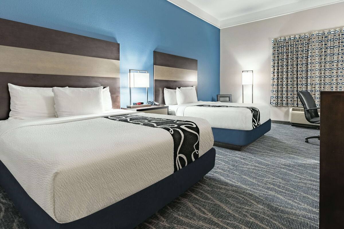 Ett hotellrum med två sängar, ett skrivbord, en stol och ett stort fönster med mönstrade gardiner.