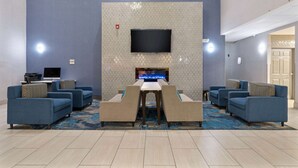 Un vestíbulo de hotel con un televisor de pantalla plana montado en una pared con textura, una mesa larga con sillas y varios sofás con cojines estampados.