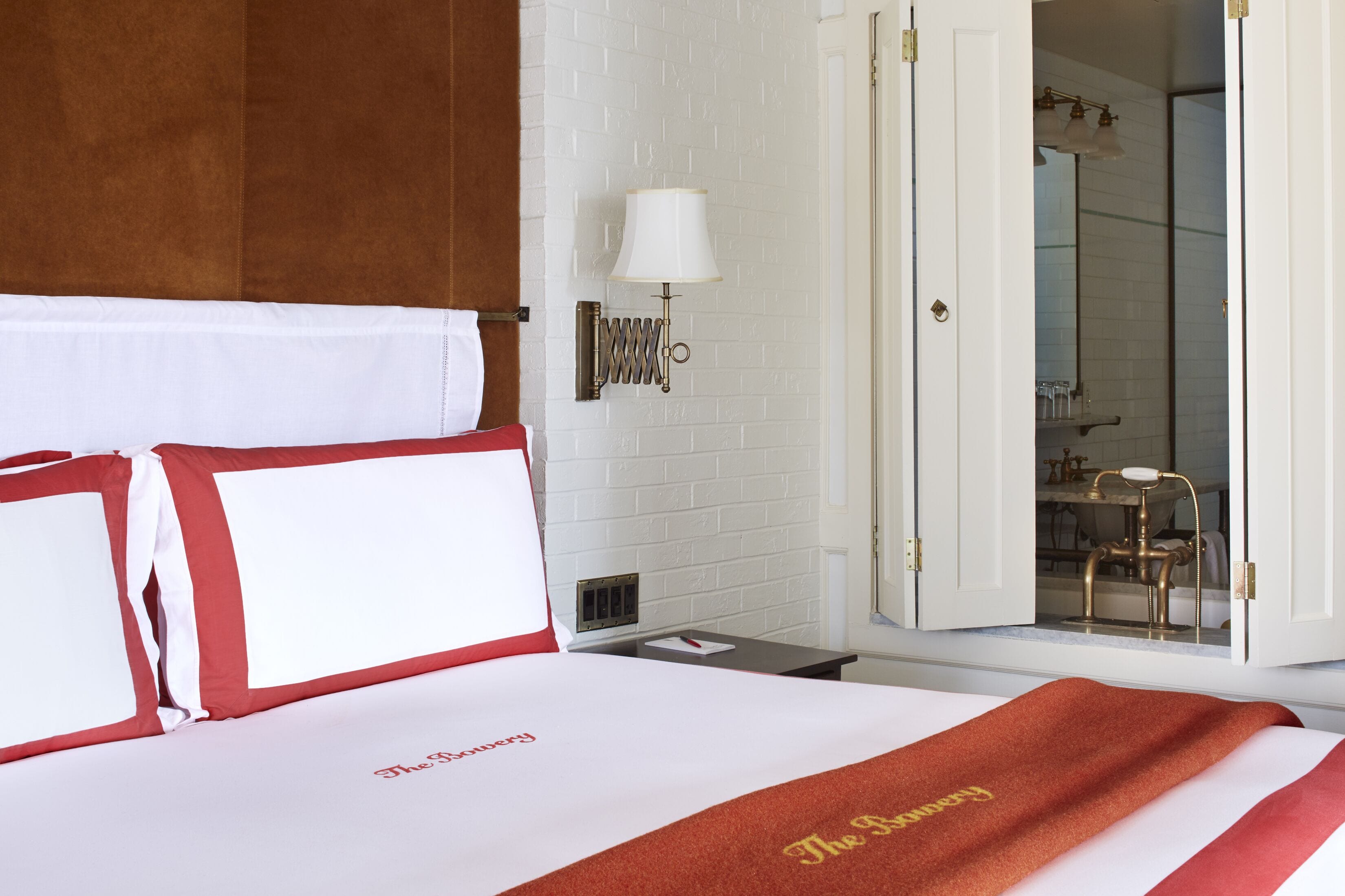Habitación de hotel con una cama bien hecha, un cojín rojo y blanco a rayas, una pared blanca con un radiador y una puerta abierta que da a un baño.