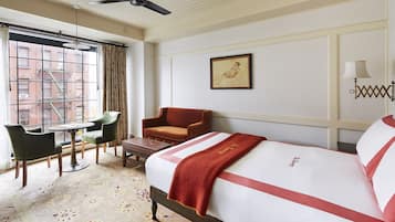Habitación de hotel con cama, una silla, una mesa y ventana con vista a edificios.