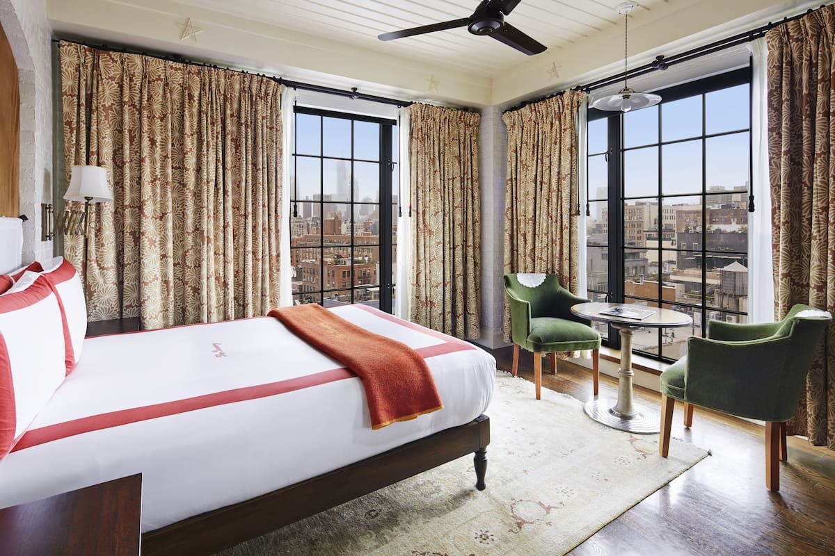 A hotel room with a large bed, two windows, a ceiling fan, a green chair, and a small table.