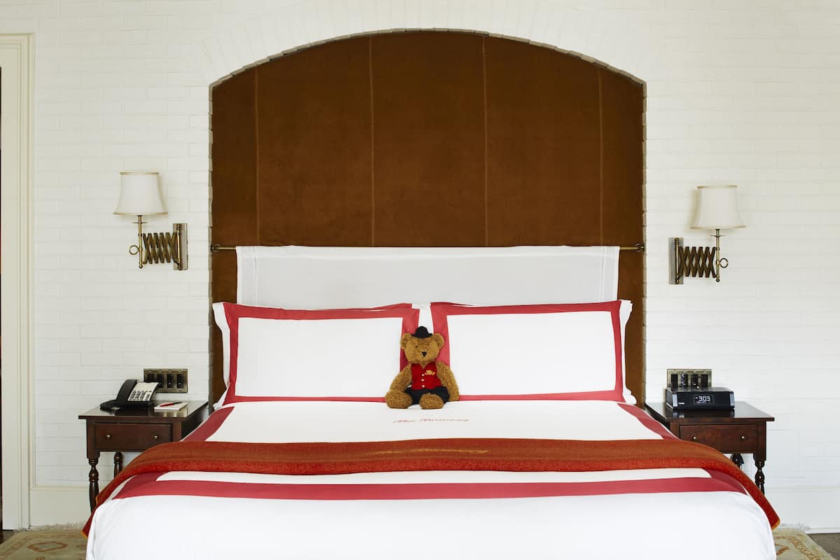 A neatly made bed with a red and white striped bedspread, a teddy bear on the pillows, and a wooden headboard. There are two bedside tables with lamps and a telephone on each.