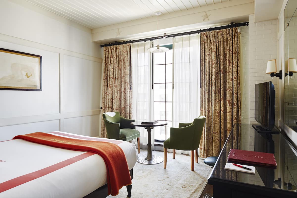 A hotel room with a bed, two chairs, a small table, a television, and a window with curtains.