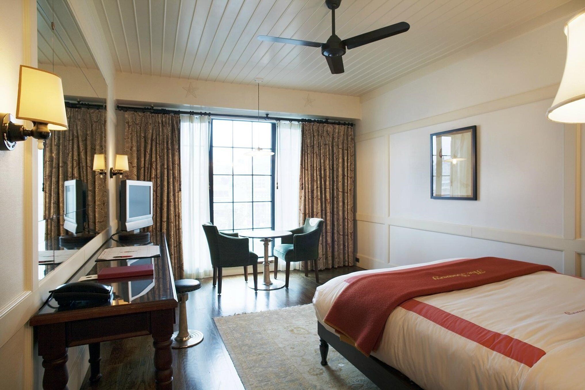 A hotel room with a bed, desk, chairs, a television, and a window with curtains.