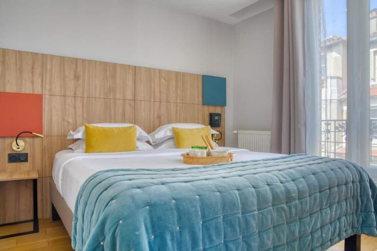 Una habitación de hotel con cabecera de madera, cama con colcha azul y vista a edificios a través de la ventana.