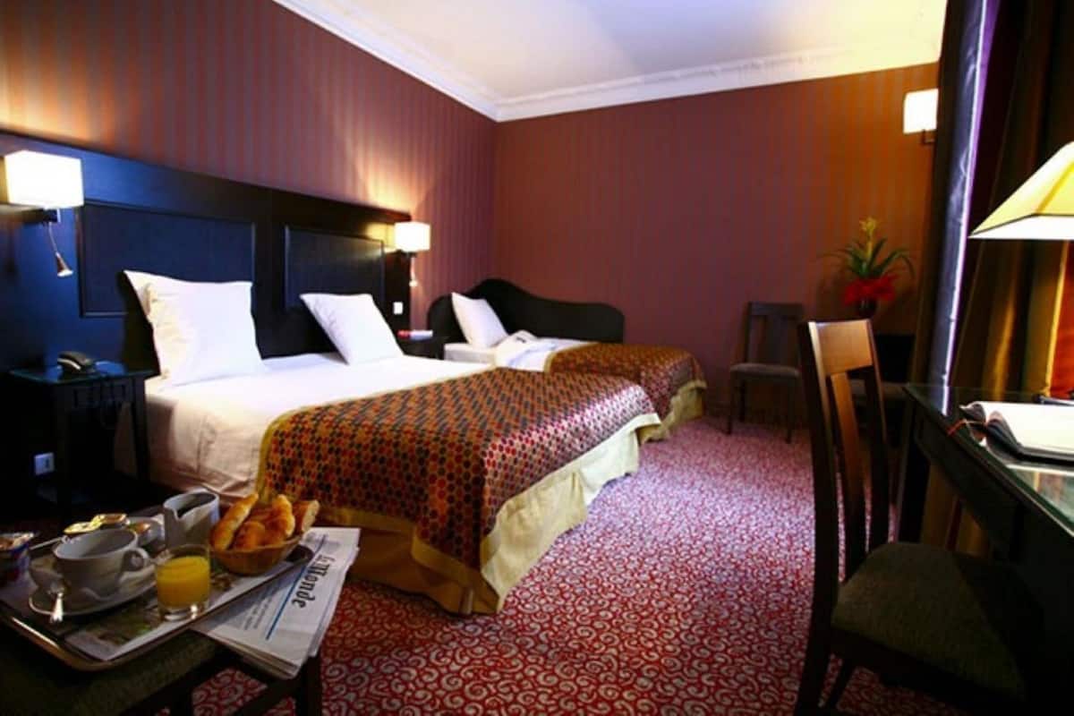 A hotel room with two beds, a desk with a chair, a small table with breakfast items, and a lamp.