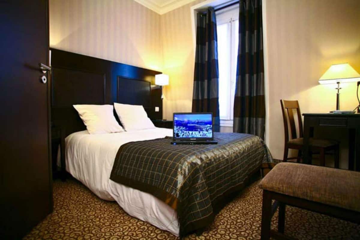 A hotel room with a bed, a laptop on the bed, a desk with a lamp, and a chair.