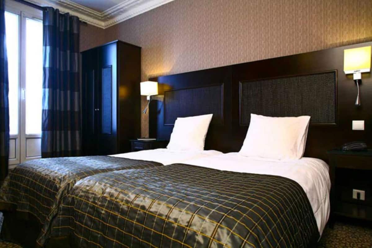 A hotel room with two beds, dark headboard, a nightstand with a lamp, and a window with curtains.