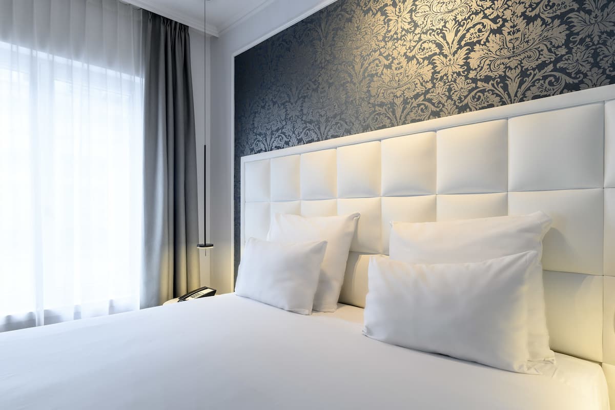 Een hotelkamer met een groot bed, wit beddengoed, een gestuwd hoofdeinde en behang met een patroon.