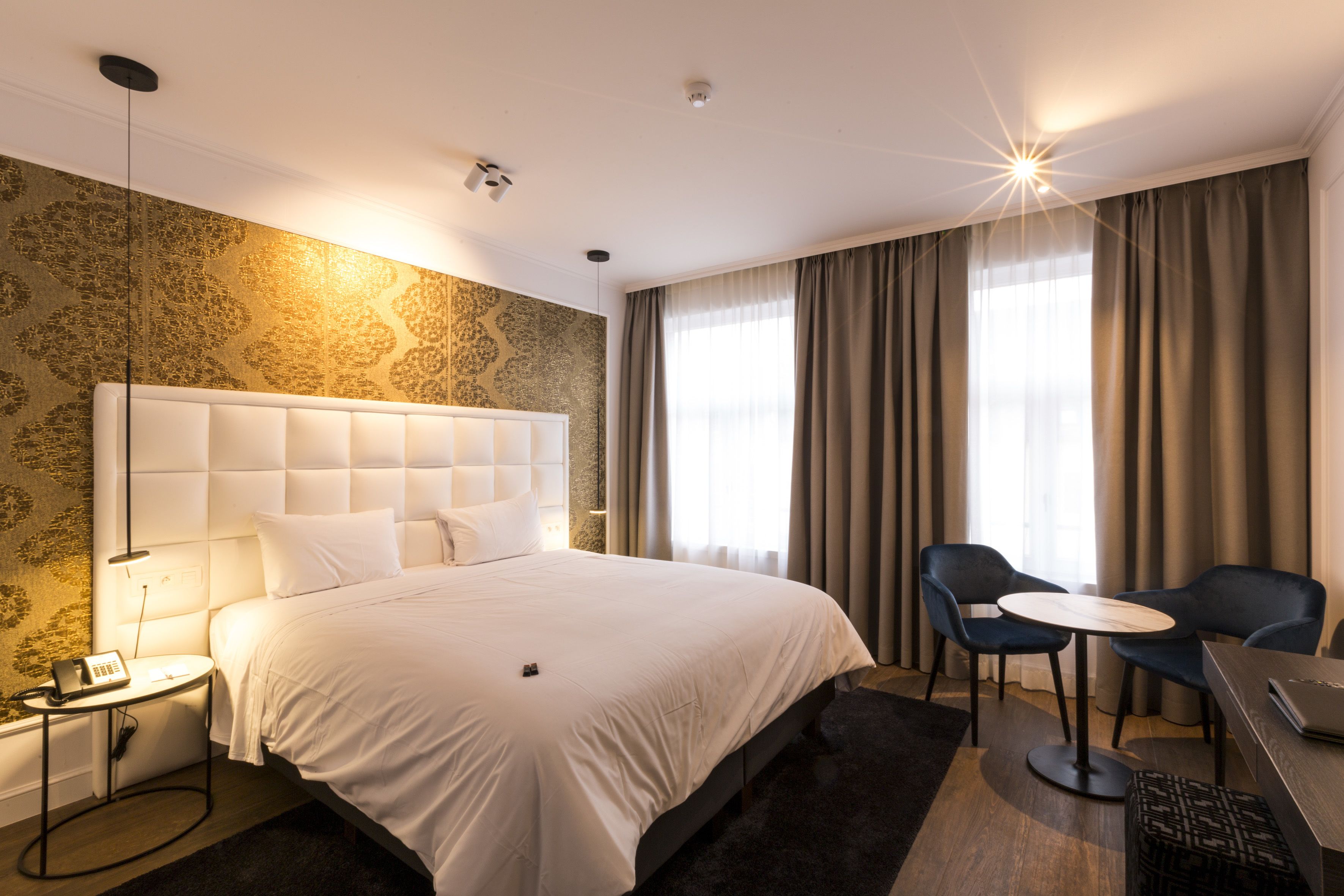 Ein Hotelzimmer mit einem großen Bett, einem Nachttisch mit Lampe, einem Sessel, einem kleinen runden Tisch und einem Schreibtisch mit Laptop.