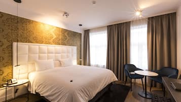 Ein Hotelzimmer mit einem großen Bett, einem Nachttisch mit Lampe, einem Sessel, einem kleinen runden Tisch und einem Schreibtisch mit Laptop.