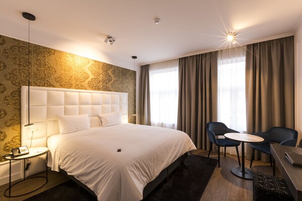 Habitación de hotel con una cama grande, una mesita de noche con lámpara, una silla, una pequeña mesa redonda y un escritorio con una computadora portátil.