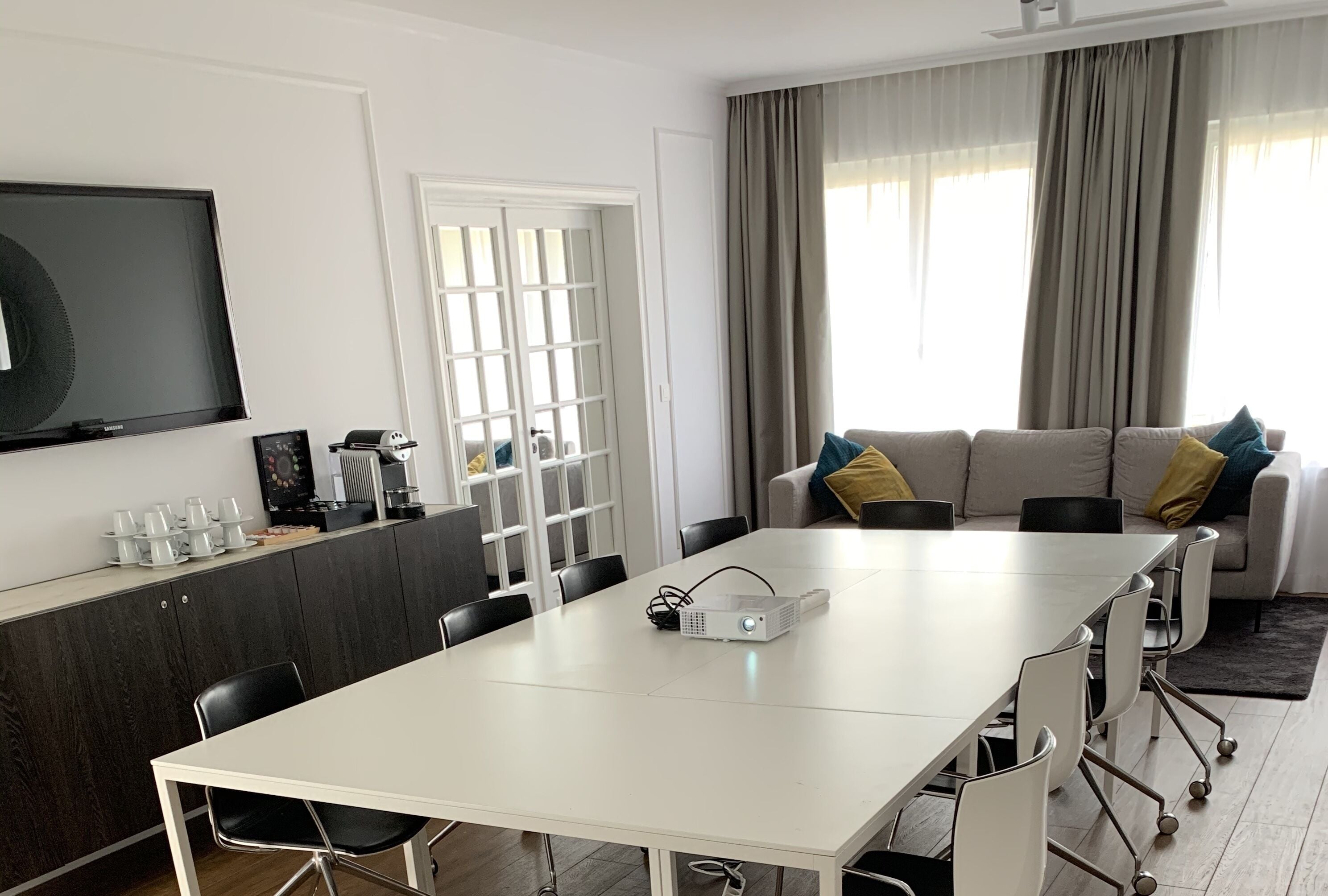A modern living room with a white dining table, black chairs, a grey sofa with yellow and blue cushions, a flat-screen TV, and a window with curtains.