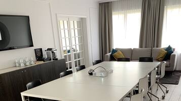 Ein modernes Wohnzimmer mit einem weißen Esstisch, schwarzen Stühlen, einem grauen Sofa mit gelb-blauen Kissen, einem Flachbildfernseher und einem Fenster mit Vorhängen.