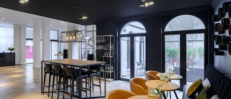 A modern interior with a wooden herringbone floor, a central dining area with a long table and chairs, and a seating area with round tables and sofas.
