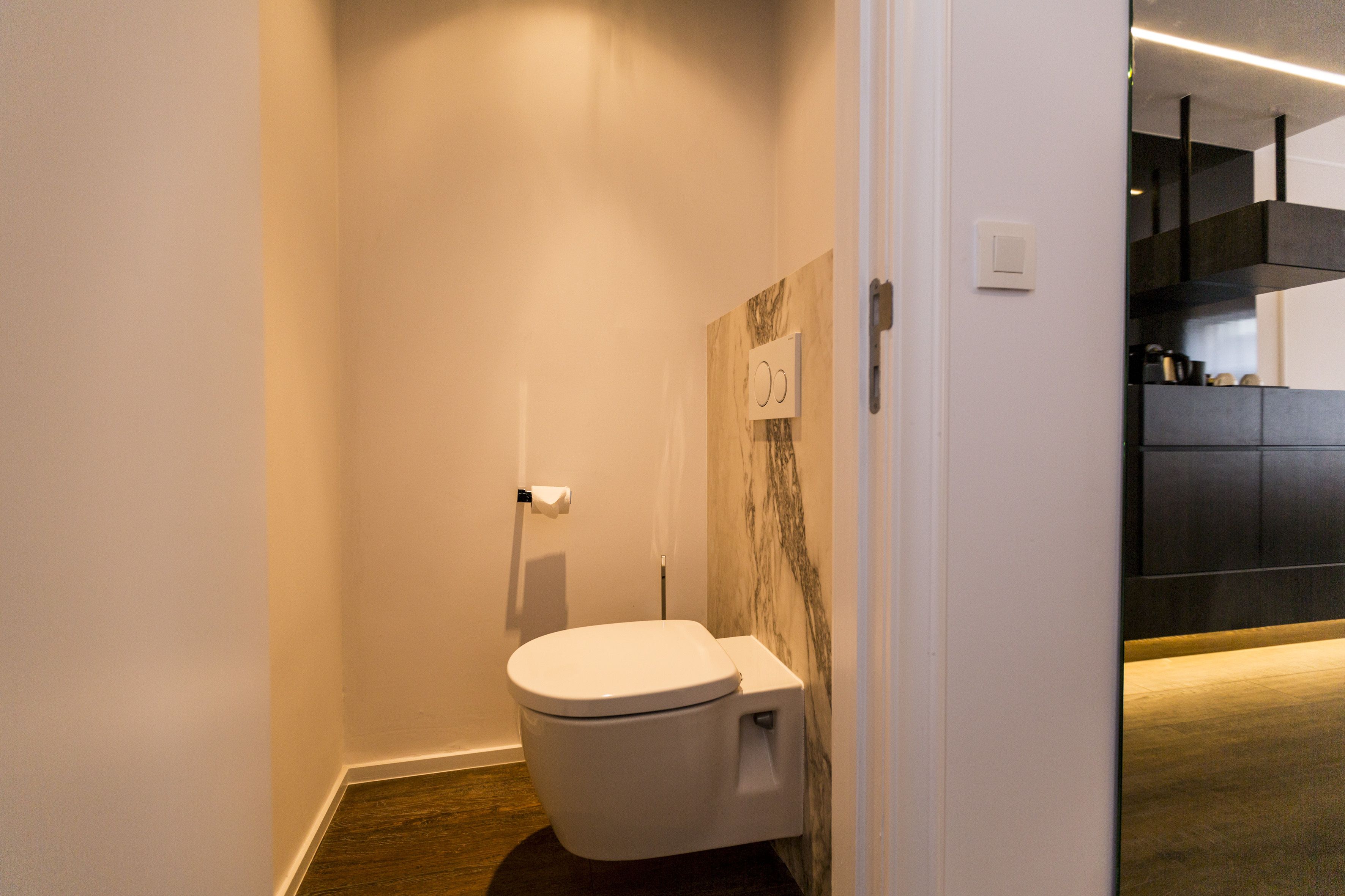 Ein modernes Badezimmer mit Toilette, einer Marmorwand und einem durch eine offene Tür einsehbarer Küchenbereich.