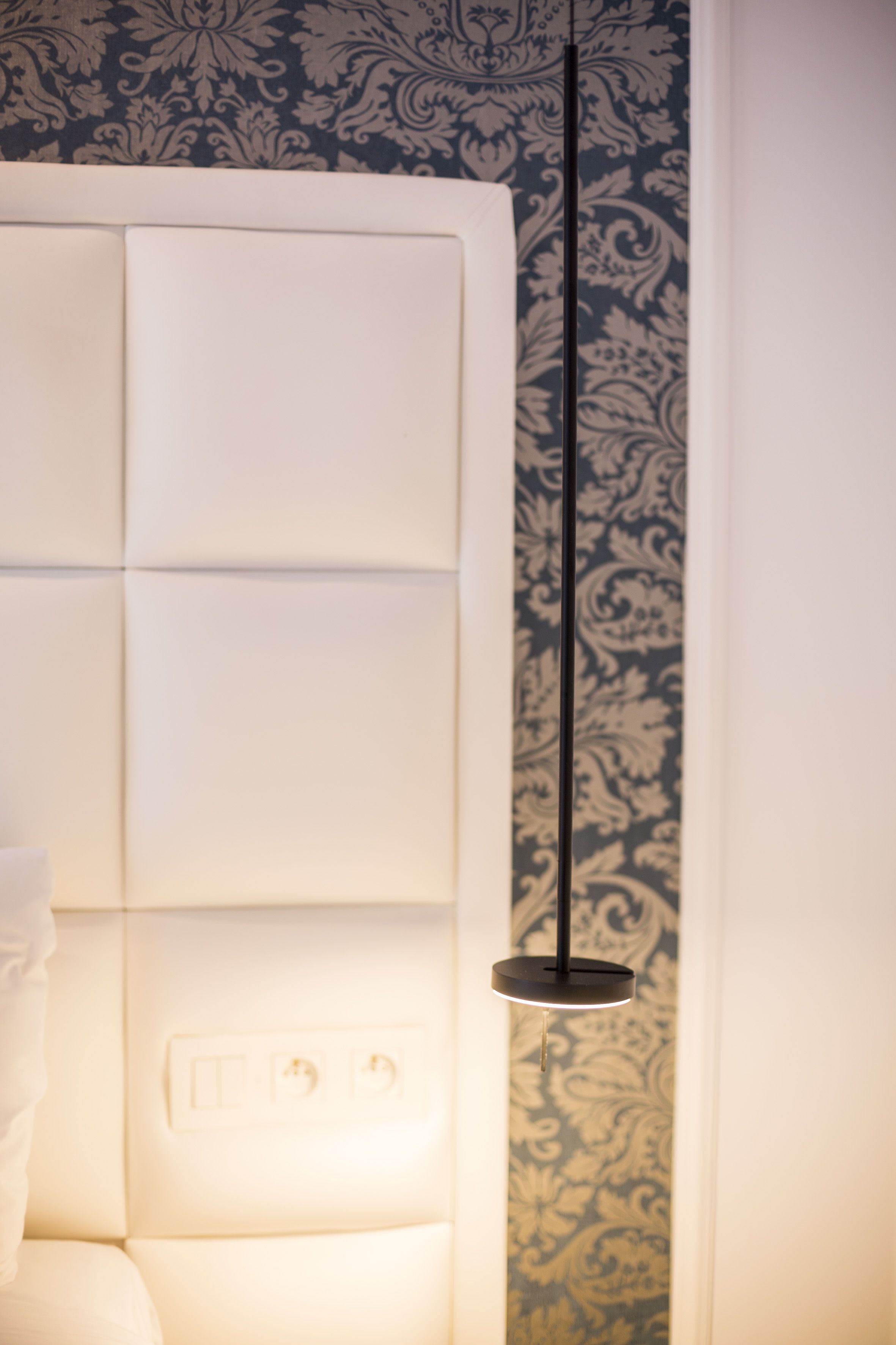 Una habitación moderna con cabecera blanca, papel tapiz estampado e iluminación colgante.