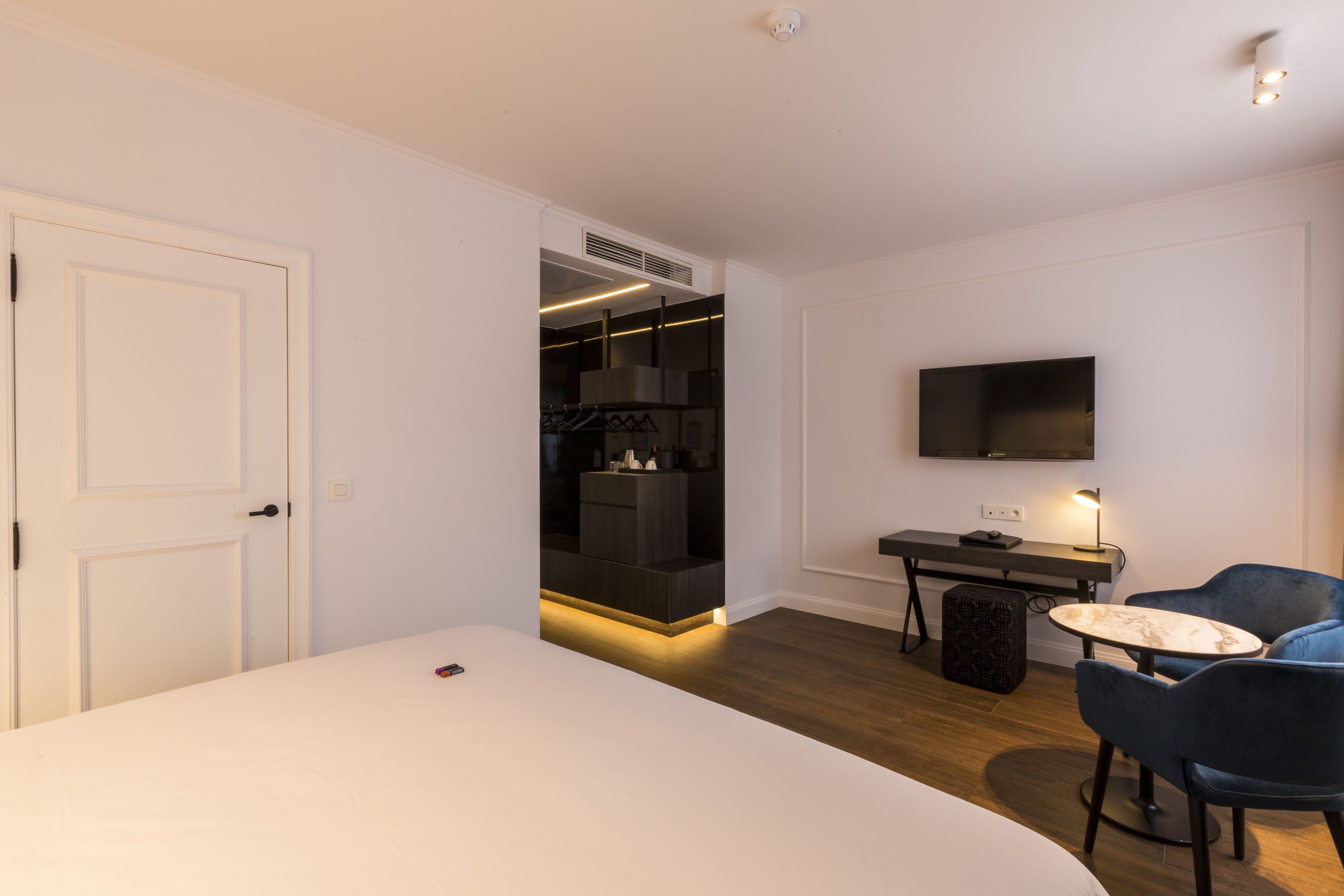A modern hotel room with a large bed, a desk with a chair, a TV mounted on the wall, and a walk-in closet.