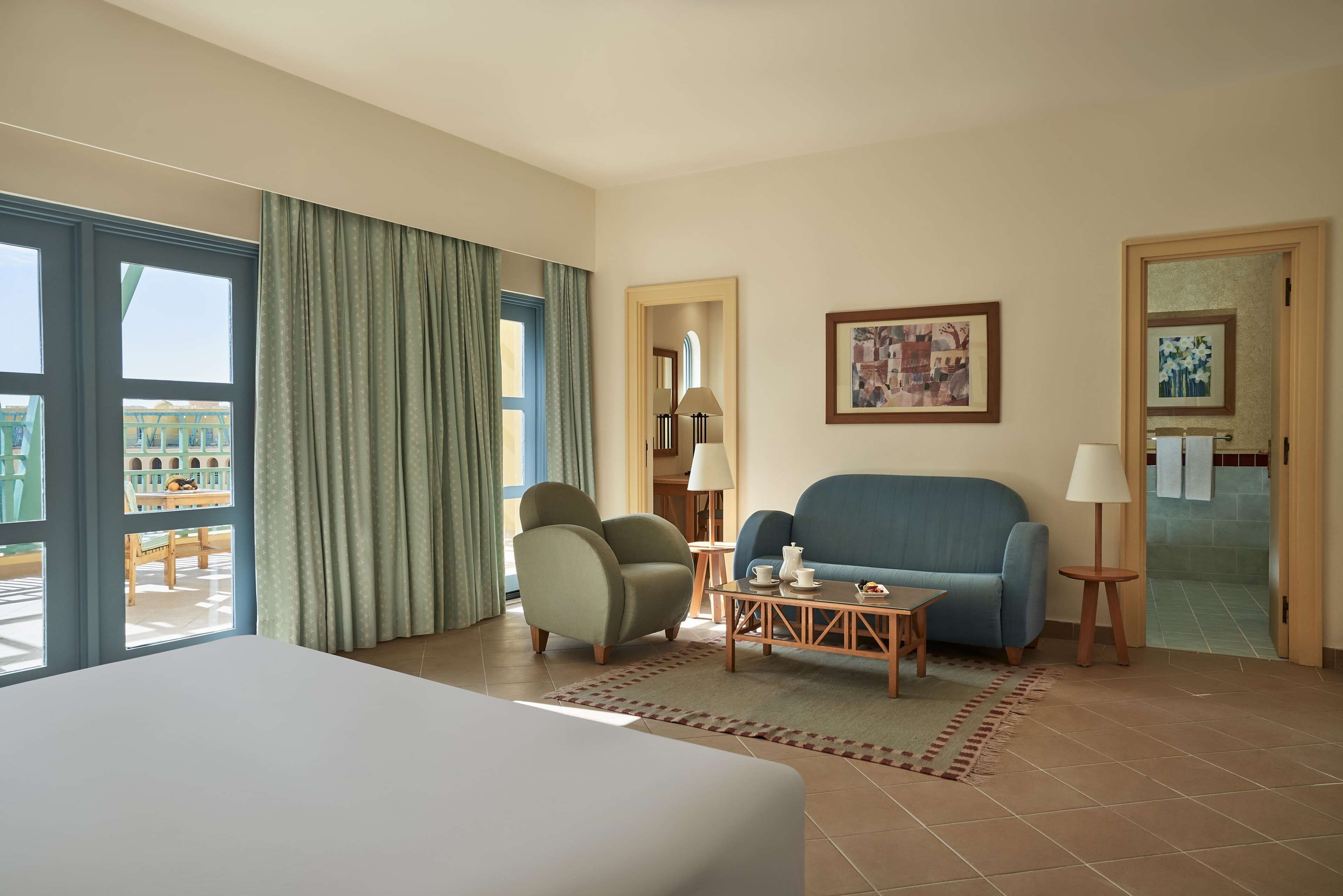 Habitación de hotel con cama, sofá azul, sillón verde, una mesita con lámpara y vista a un edificio a través de la ventana.