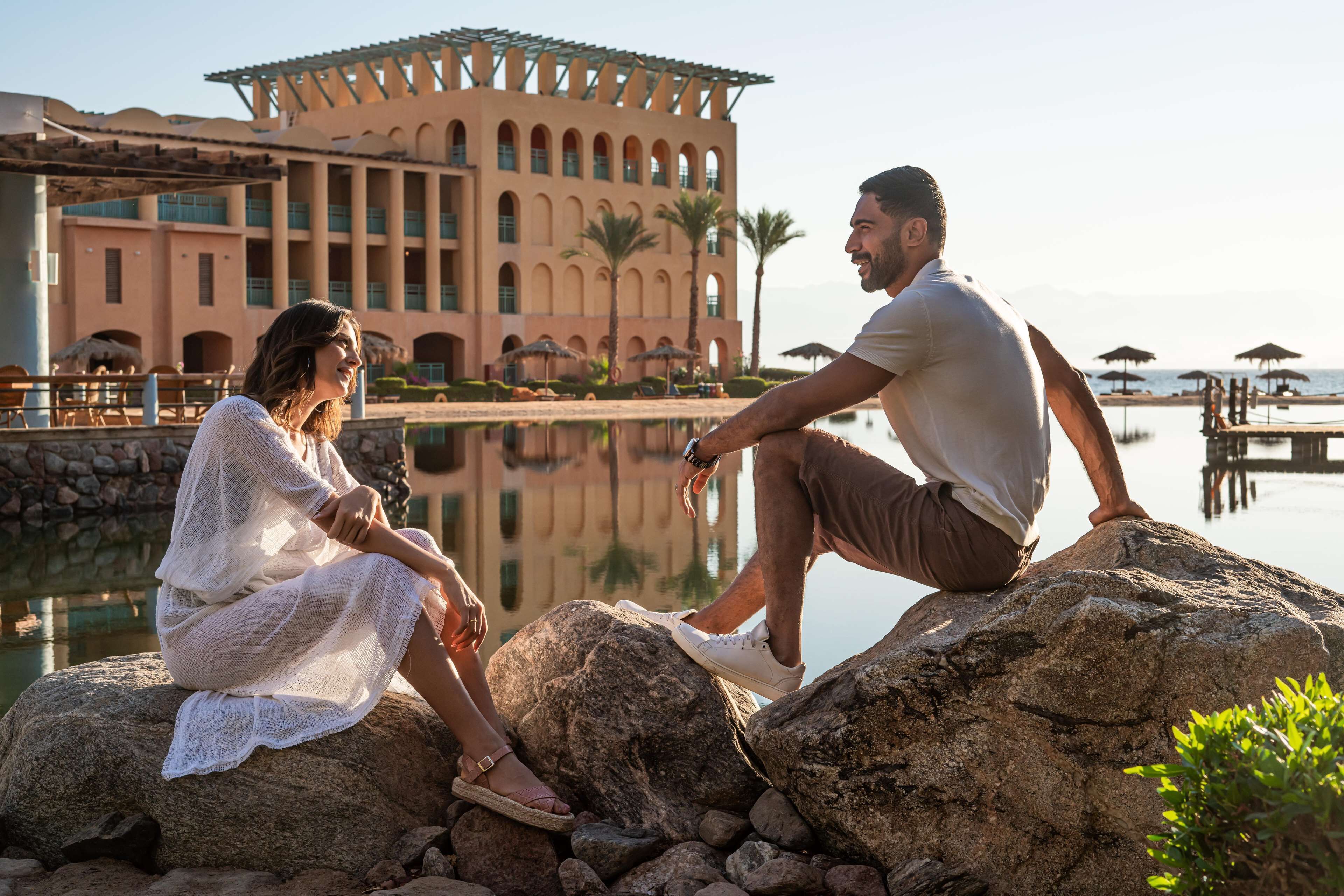 A couple sitting on rocks by a reflective pool with a large building in the background.