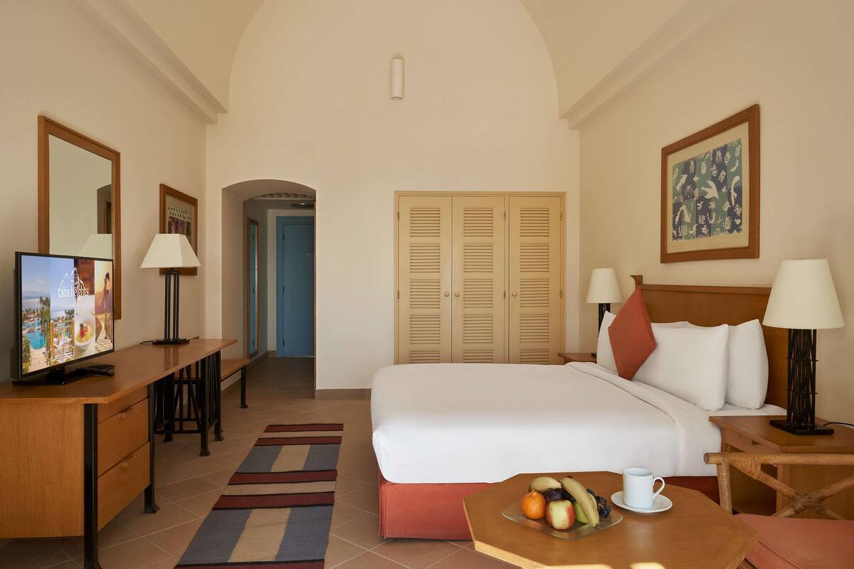 Una habitación de hotel con cama, escritorio con televisor, mesa de comedor con fruta y un cuadro en la pared.
