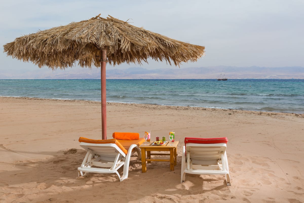 Una playa con dos sillas de playa blancas, una mesa con bebidas y un quiosco de paja.
