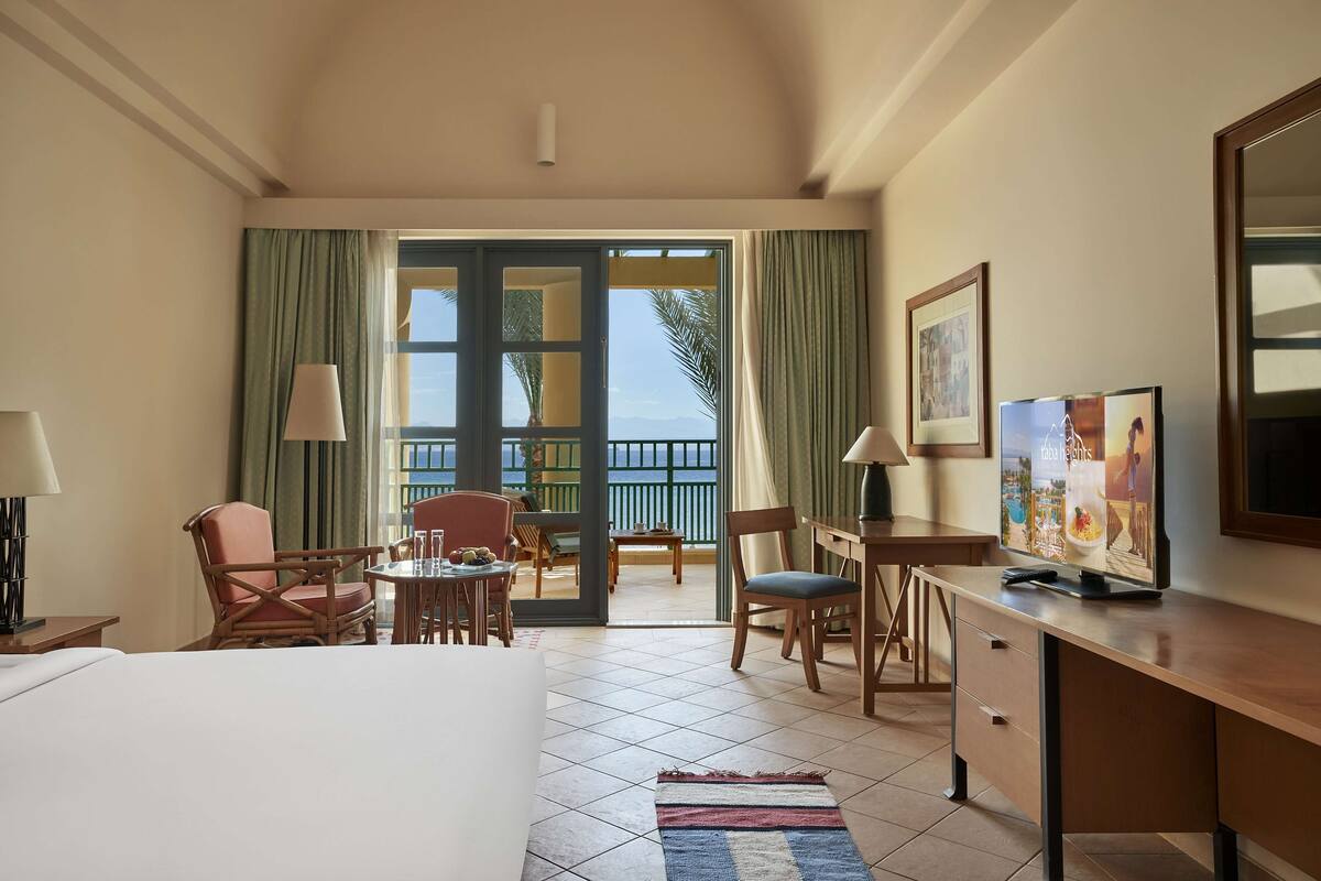 Habitación de hotel con cama, escritorio, sillas y vista al mar a través de amplias ventanas.