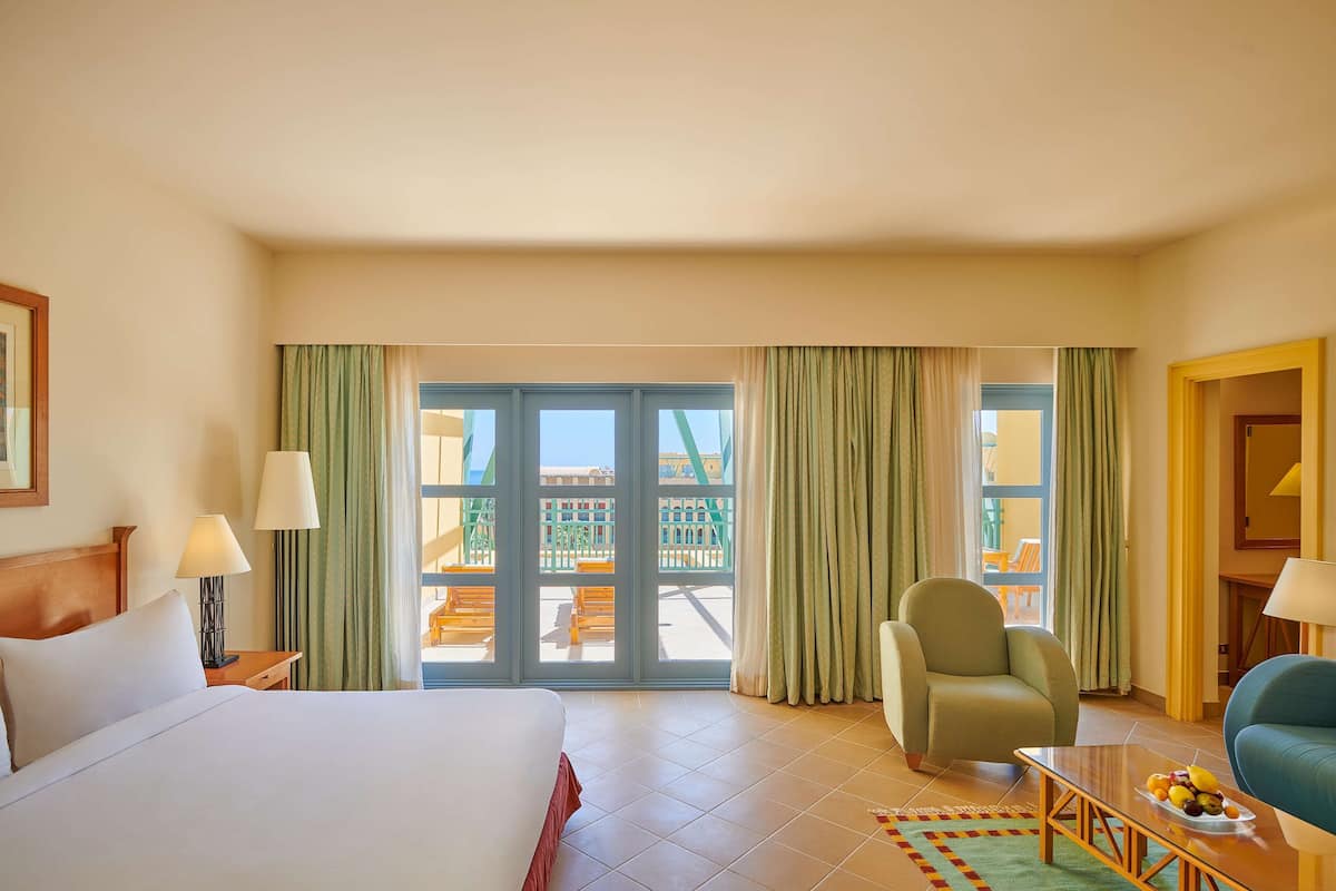 Una habitación de hotel con una cama, dos sillas, una mesa con fruta y un balcón con cortinas.